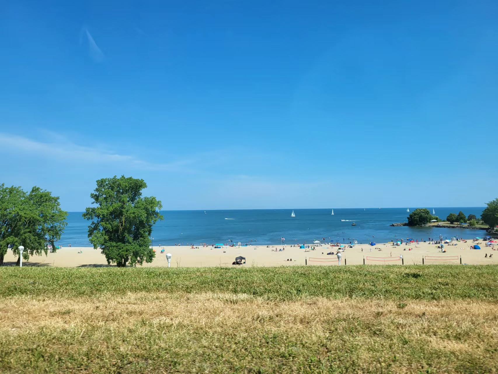 A picture of a beach with people resting on the sand. There are a few trees on the hill where the photo was taken, and sail boats on the water.