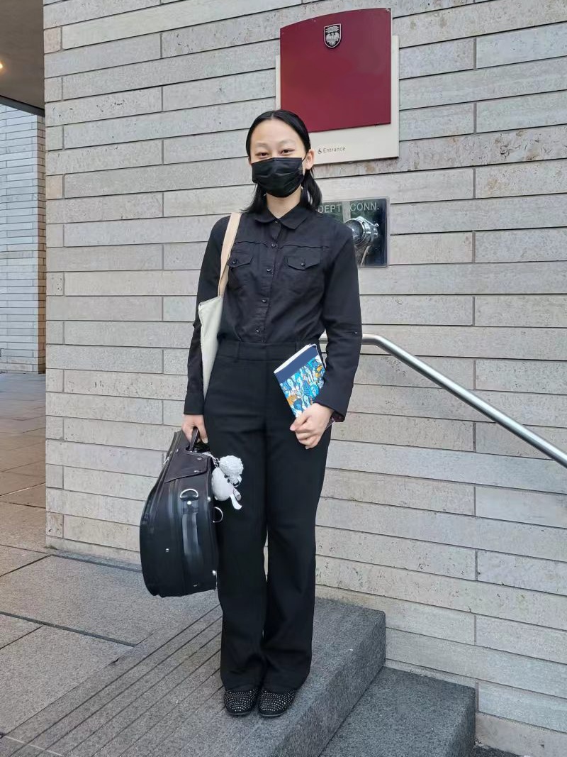 A picture of Grace wearing full black, carrying their violin case and a concert booklet. They stand in front of a white brick building.