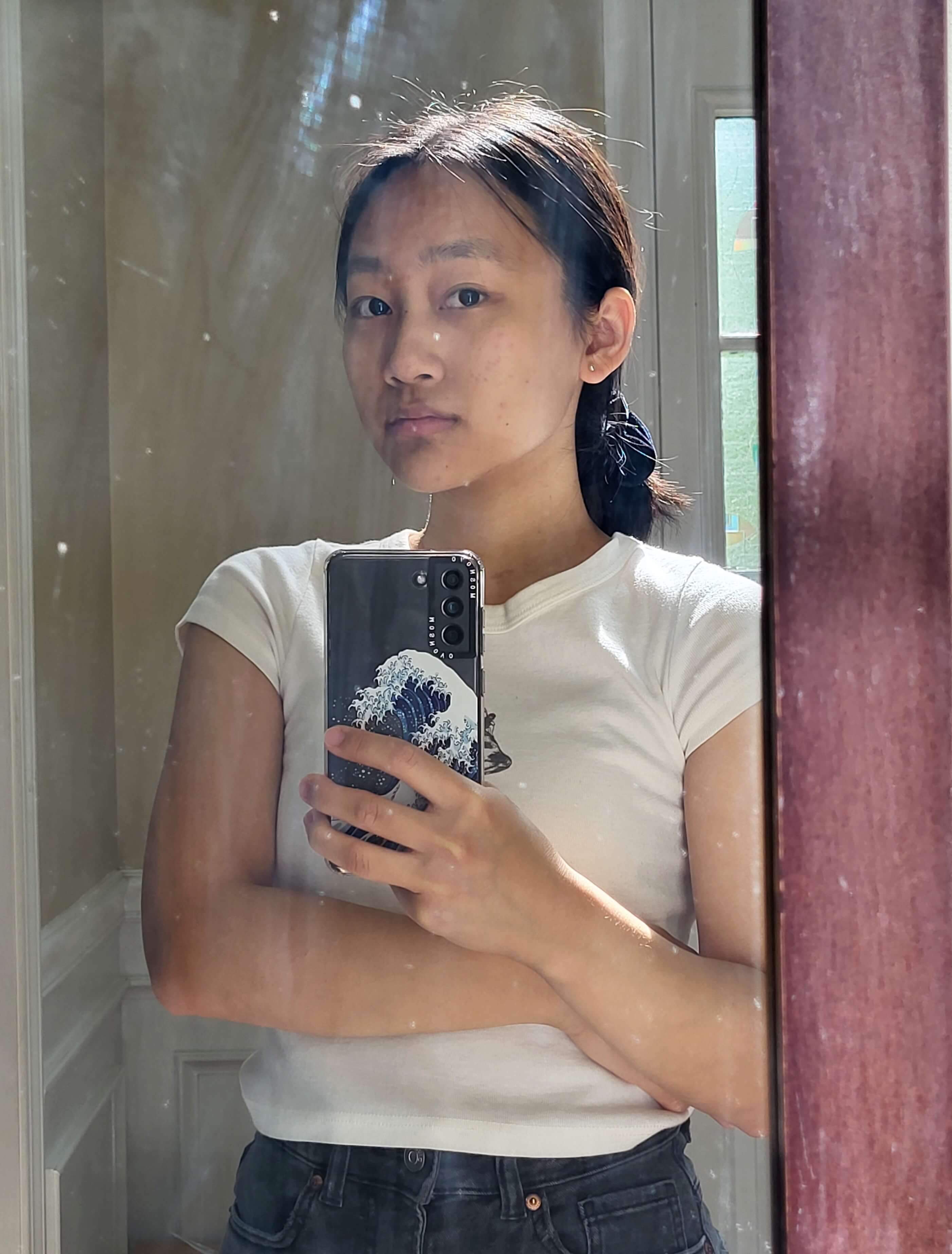 A picture of Grace in the mirror. They have a neutral face and stands with one arm crossed.