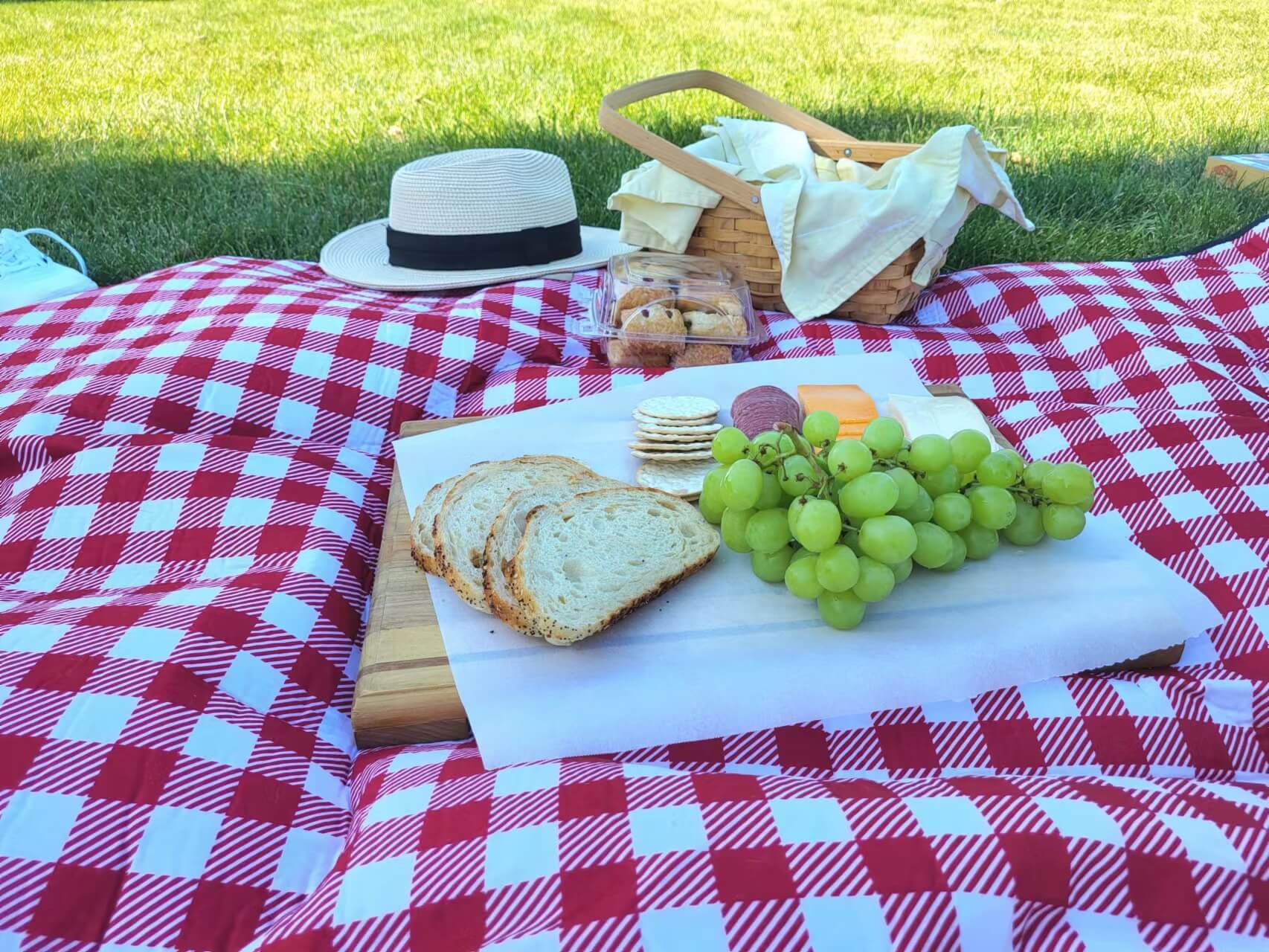 A picnic blanket with a wooden board that has bread, grapes, crackers, cheese, and salami on it. In the back, there is a basket, a hat, and a pair of white Vans.