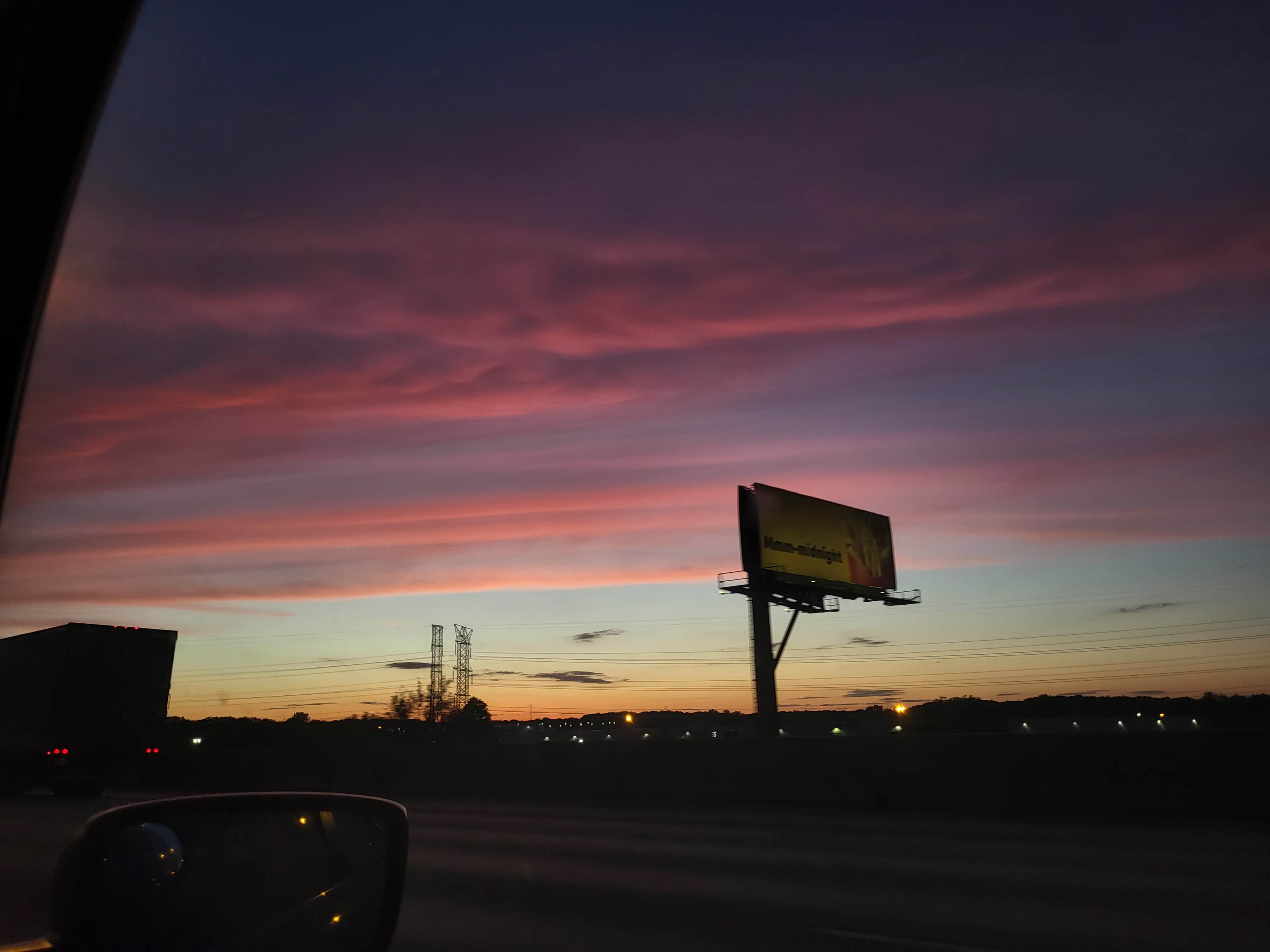 Through the car window, the photo captures a sunset consisting of pink and orange colors. The sky is cloudy.